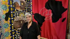 Uma mulher com cabelos grisalhos em pé. Ao fundo dela há um mural com um desenho do mapa da África ligado ao América do Sul