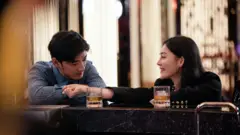 Uma imagem do jogo *Emotional Anti-Fraud Simulator* (Simulador Emocional Anti-Golpes) mostra um homem e uma mulher conversando sentados em um bar, com dois copos de bebida à frente. O homem veste uma camisa azul de manga longa, e a mulher usa um cardigã preto e brincos dourados.