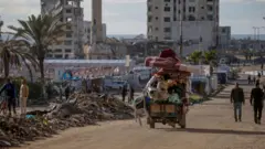 فلسطينيون يصلون إلى وسط مدينة غزة بعد نزوحهم من شمالي القطاع، يصلون مشياً أو في عربة قديمة. الصورة التقطت نهاراً قرب خيام ودمار حول الطريق.