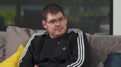 A imagem mostra Casimiro Miguel sentado em um sofá durante uma entrevista, usando um moletom preto com listras brancas nas mangas e um microfone de lapela preso à roupa. Ela olha para alguém fora do enquadramento, em um ambiente interno com fundo desfocado.