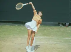 Fotografía a color con Bueno saltando con raqueta en alto, con un vestido con encaje blanco y falda plizada, braga de encaje y un sweater amarillo apagado, sobre pista de grama.