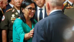 Delcy Rodriguez is wearing a green suit and glasses while greeting greets aa man whose back is toward the camera. sShe has her hand on his arm and is smiling. A man in uniform and a crow is behind her