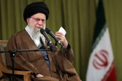 Ali Khamenei falando em um microfone. Ele está sentado em uma cadeira e ao fundo há uma bandeira do Irã