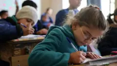 در تصویر دختربچه‌ای پشت میز نیمکت مدرسه در حال نوشتن مشق با یک خودکار آبی دیده می‌شود.