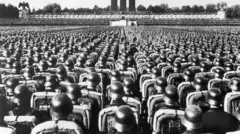 Foto em preto e branco mostra milhares de soldados com capacetes pretos, voltados para um grande palco com três grandes colunas. O Partido Nazista realizava comícios semelhantes aqui todos os anos entre 1933 e 1938.