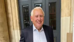 Sir Bob Neill está em frente a um edifício com grandes portas cinza-escuras e paredes de pedra bege. Ele está vestido com um paletó escuro sobre uma camisa xadrez azul-clara de gola aberta.