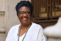 Rosana Paulino é uma mulher negra. Ela usa óculos de grau com armação preta, uma faixa preta no cabelo curto e uma blusa branca com um colar.