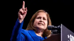 Hindistonlik 61 yoshli G’azala Hoshimiy ko‘k kostyumda, yelkasigacha bo‘lgan qora sochli, minbarda mikrofonga gapirmoqda. Uning o‘ng qo‘li yuqoriga ko‘tarilib, ishora barmog’i yuqoriga ko‘rsatilgan.