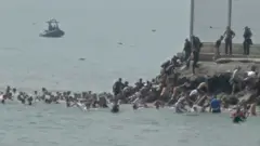 Dezenas de pessoas estão no mar enquanto outras sobem uma pequena encosta para chegar à terra firme. Pessoas com uniformes de segurança observam a cena às margens. Uma pessoa estende a mão enquanto migrantes caminham em direção aos que sobem a encosta. Ao fundo, há um pequeno barco no mar.