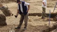 Dave Derby con su detector de metales en la granja Whitehall durante la excavación de 2003. Está inclinado mirando al suelo y sosteniendo el detector con la mano derecha. Detrás de él se ven zanjas y terreno despejado.