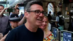 Andy Burnham, ladeado por policiais. Há multidões de pessoas tirando fotos atrás dele. Ele usa óculos e uma camisa escura.