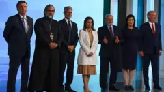 Foto em um estúdio de TV com Bolsonaro, padre Kelmon, Felipe d'Avila, Soraya Thronicke, Lula, Simone Tebet e Ciro Gomes, da esquerda para a direita.