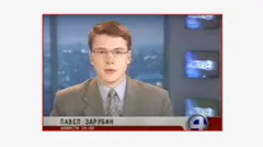 Un joven Pavel Zarubin presenta un noticiero en un canal de televisión ruso con el logo "4" en la esquina inferior derecha de la pantalla. Zarubin viste traje gris, corbata gris y lentes. Las pantallas de televisión están dispuestas verticalmente en un panel a la derecha de la pantalla. El fondo es el horizonte de una ciudad.