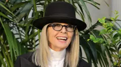 Diane Keaton luce un sombrero y un traje negro elegante y sonríe mirando a un costado rodeada de plantas ornamentales.