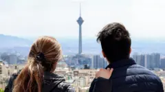 زن و مردی از بالای بلندی به نمایی از تهران که برج میلاد در آن مشخص است نگاه می کنند