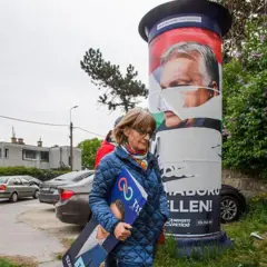 Orban'In yırtılmış bir posterinin yanından geçen gözlüklü ve mavi ceketli bir kadın elinde yüzü görülmeyen başka bir siyasetçinin afişini tutuyor