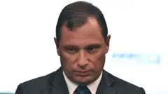 Mauro Cid com olhar consternado, olhando levemente para baixo em frente a fundo branco