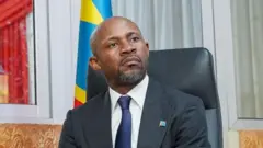 Patrick Muyayayambaye ikoti n'ikaruvati vyirabura, inyuma yiwe hari ibendera ya DRC