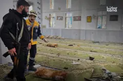 صورة تظهر آثار الانفجار داخل مسجد الإمام علي بن أبي طالب في حي وادي الذهب في حمص. يقف رجلا أمن في المسجد.
