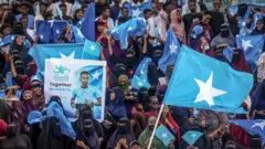 زنانی با روبنده در جلوی تصویر و مردانی در پشت با پرچم‌های سومالی و پوستری از عمر عرتن در تصویر هستند.