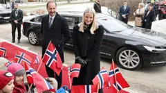Una pareja vestida con abrigos negros está de pie delante de un coche, mientras unos niños en primer plano agitan banderas de Noruega.