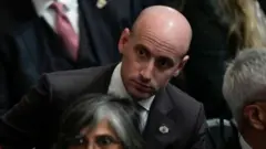 Stephen Miller, sentado, olha de lado para observar algo com atenção