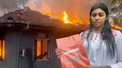 ගිනි ගන්නා නිවසක් ඉදිරිපිට සුදු පාට බ්ලවුස් එකක් ඇඳගත් තරුණියක් රතු පාට පොතක් අතේ තබාගෙන සිටින අයුරු පෙනේ.