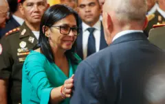 Delcy Rodriguez is wearing a green suit and glasses while greeting greets aa man whose back is toward the camera. sShe has her hand on his arm and is smiling. A man in uniform and a crow is behind her