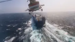 Lado de baixo de um helicóptero houthi se aproximando por trás do enorme navio de carga Galaxy Leader, que deixa uma imensa onda para trás ao viajar em mar aberto, sem terra à vista