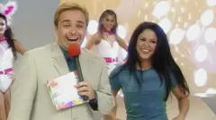 O apresentador Gugu, de terno claro e microfone vermelho, sorri para a câmera ao lado de Shakira, de roupa azul. Ao fundo, dançarinas vestidas de branco com detalhes em rosa aparecem em um estúdio iluminado do programa de auditório Domingo Legal.
