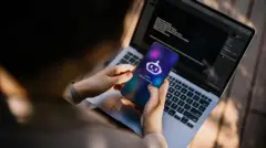 Uma pessoa segura um telefone com um chatbot de IA na tela dizendo, em inglês: "Olá! Pergunte-me qualquer coisa...". Abaixo, há um laptop com tela preta e texto branco.