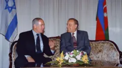 Aliyev ve Netanyahu koltukta gülümseyerek sohbet ediyor. Arkada İsrail ve Azerbaycan bayrakları, önlerindeki masada bir buket çiçek var.