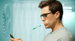 Homem de perfil no banheiro, usando óculos com uma escova de dentes na boca, enquanto consulta seu telefone celular