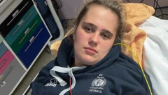 Phoebe Tesoriere deitada em uma cama de hospital, com agasalho azul