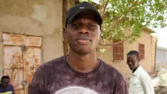 Debout à l'ombre d'un arbre, un jeune homme portant une casquette de base-ball noire regarde droit dans la caméra.