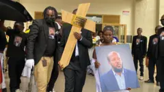 Umukobwa ateruye ifoto ya Aimable Karasira n'undi musore ateruye umusaraba uriho amazina ya Karasira n'umwirondoro we