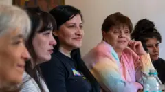 Cinco mulheres de diferentes idades estão sentadas à mesa, voltadas para a mesma direção. Andrea, a quarta da esquerda para a direita, veste um casaco em tom pastel e leva a mão ao rosto.