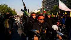 سيدة إيرنية ترتدي ملابس سوداء وعصابة رأس حمراء وتحمل بندقية آلية تركب دراجة نارية خلف شخص يرتدي نفس الملابس وحولهم حشود ترتدي ملابس سوداء وترفع أسلحة أثناء مسيرة في العاصمة طهران رفضاً للهجمات الأمريكية الإسرائيلية