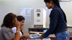 Duas mesárias atendem eleitora em local de votação com a cabina de votação ao fundo