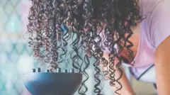 Uma pessoa usa um secador de cabelo com um difusor grande para secar os cabelos cacheados. A cabeça da pessoa está inclinada para a frente, de modo que os longos cachos escuros pendem em direção ao difusor.
