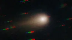 Комета 3I/Atlas показана в виде яркой точки, окруженной туманной сферой и хвостом слева. На заднем плане видны полоски синих, зеленых и красных точек, представляющие звезды, которые движутся относительно кометы.