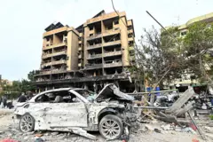 Carro branco seriamente danificado em frente a um edifício fortemente atingido, rodeado por destroços