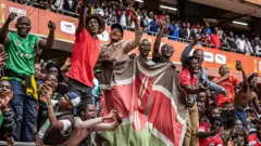Des supporters brandissent un drapeau kenyan tandis qu'un groupe de personnes crie des encouragements dans une tribune bondée du stade Kasarani lors d'un match entre le Kenya et le Maroc au Championnat d'Afrique des Nations 2024.