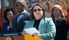 Kathleen Corradi, com outras pessoas ao lado, em um palanque, com um papel na mão