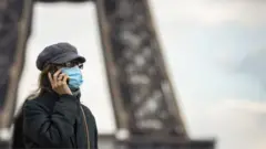 A woman wearing a surgical face mask walks near the Eiffel Tower