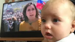 Bebé Isadora con mamá Katy Watson en la televisión detrás de ella
