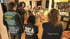 Fotografia colorida mostra policiais de pé em um apartamento cheio de documentos e pessoas
