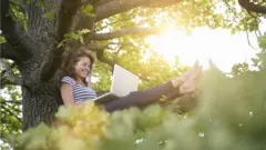 A woman working on a laptop sits in the branches of a tree, barefoot, surrounded by greenery illuminated by the summer sun