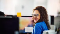 Mulher de óculos e casaco azul ri no escritório