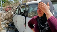 Rima Zahed stands near a damaged car at the entrance of the port of Beirut as she awaits for news of her missing brother Mohammed, a port employee, on August 8, 2020.